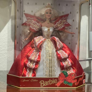 May include: A red and white Barbie doll wearing a crown and a red and white gown with gold accents. The doll is in a clear plastic box with a red and gold background. The box says "Happy Holidays" and "Special Edition Barbie".