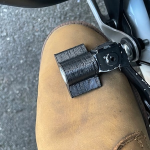 以下が含まれることがあります： 黒色の3Dプリントされたオートバイ部品が黒色のレバーに取り付けられ、茶色の革ブーツの上に置かれています。ブーツには茶色の靴紐があり、使い古された外観です。背景にはオートバイの一部と灰色の表面が含まれています。