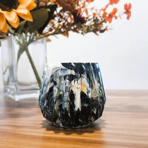 Puede incluir: Un jarrón decorativo facetado con un patrón veteado en negro, blanco y dorado. El jarrón está sobre una superficie de madera, con un jarrón de vidrio transparente y flores en el fondo. Decoración del hogar.