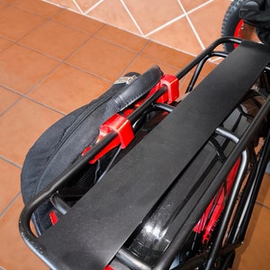 Puede incluir: Un portaequipajes negro para bicicleta con una parte superior plana y soportes curvos. Piezas de plástico rojo aseguran el portaequipajes a la bicicleta. Una bolsa negra está sujeta al portaequipajes. La bicicleta tiene neumáticos rojos.