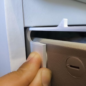 Puede incluir: Una mano interactúa con un componente de plástico blanco, posiblemente un pestillo o un clip, dentro de un armario o compartimento de color claro. El componente es rectangular con un borde texturizado. El fondo muestra una superficie beige con una hendidura circular.