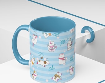 Può includere: Tazza in ceramica con interno, manico e bordo blu. L'esterno presenta un motivo a righe azzurre con gatti dei cartoni animati in varie pose, con sciarpe e cappelli. La tazza è alta circa 10 cm.