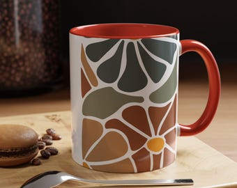 May include: A white ceramic mug with a red interior and handle. The mug has a floral design in brown, green, and yellow. A spoon and a macaron are on a wooden surface beside the mug.