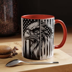 Könnte beinhalten: Eine weiße Keramiktasse mit rotem Innenraum und Henkel. Die Tasse zeigt eine schwarz-weiße Grafik eines Adlerkopfes vor dem Hintergrund der amerikanischen Flagge. Die Tasse steht auf einer Holzoberfläche.