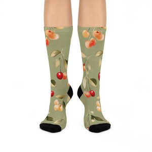 May include: Knee-high socks featuring a light green backdrop adorned with a cherry and leaf pattern. The cherries are red and yellow, with green leaves. The socks have black heels and toes.