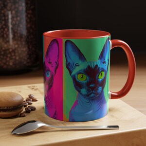 May include: A colourful ceramic mug with a pop art style Sphynx cat design. The mug has a red interior and handle, with the cat's face in blue, green, pink, and purple. The mug is on a wooden surface, with a spoon and a macaron.
