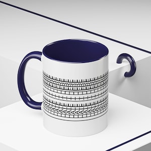 May include: White ceramic mug with a navy blue interior and handle. The mug features a black geometric pattern around the exterior. The mug is sitting on a white surface.
