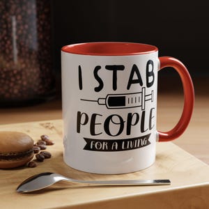 May include: White ceramic mug with a red interior and handle. The mug features the text "I STAB PEOPLE FOR A LIVING" in black, along with a syringe graphic. A spoon and a macaron are on a wooden surface.