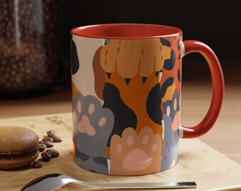 Könnte beinhalten: Eine Keramiktasse mit rotem Innenraum und Henkel, verziert mit einem sich wiederholenden Muster aus bunten Pfotenabdrücken in Grau-, Beige- und Orangetönen. Die Tasse steht auf einer Holzoberfläche mit einem Löffel und einem Gebäck.