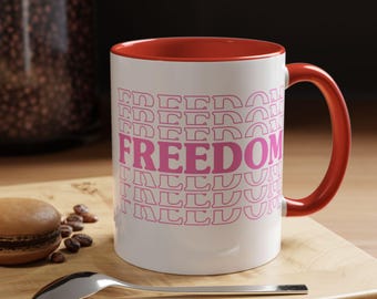 May include: White ceramic mug with a red interior and handle. The mug features the word "FREEDOM" in pink, with a repeating pattern. A spoon and a pastry are on a wooden surface.