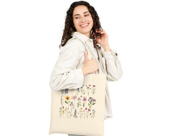 May include: A cream-colored tote bag featuring a watercolor illustration of various wildflowers in shades of purple, yellow, and pink. The bag is held by a person wearing a white button-down shirt and blue jeans.