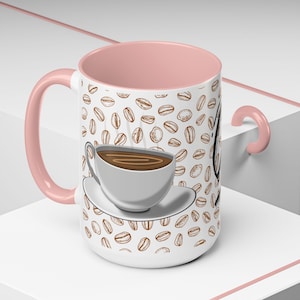 Könnte beinhalten: Weißer Kaffeebecher mit rosa Innenraum und Griff. Der Becher ist mit braunen Kaffeebohnen-Illustrationen und einer Grafik einer Kaffeetasse und Untertasse verziert. Der Becher steht auf einer weißen Oberfläche.
