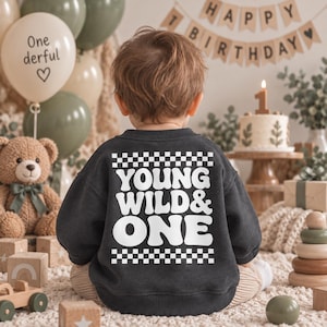 Könnte beinhalten: Ein dunkelgraues Langarm-Sweatshirt mit weißem Schriftzug YOUNG WILD & ONE auf der Rückseite, eingerahmt von einem Schachbrettmuster oben und unten. Das Kleidungsstück wird von einem Kind von hinten in einer Geburtstagskulisse getragen.