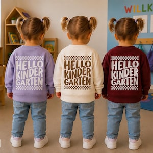 Könnte beinhalten: Drei Kinder von hinten gesehen, bekleidet mit hellblauen Jeans und langärmeligen Sweatshirts in Lavendel, Creme und Kastanienbraun. Jedes Sweatshirt hat einen weiß karierten Rand und den Text HELLO KINDER GARTEN in welliger Schrift.