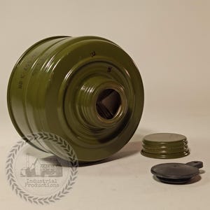 Puede incluir: Un filtro de máscara antigás verde oliva con un cuerpo cilíndrico y una abertura central. Dos tapas, una verde oliva y otra negra, se colocan junto al filtro. El texto "Industrial Productions" es visible en la parte inferior izquierda.