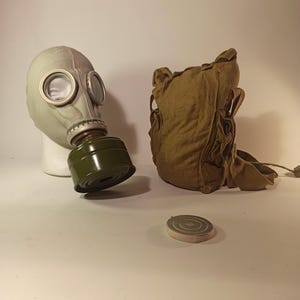 May include: A vintage gas mask with a light grey rubber facepiece, clear eye lenses, and a dark green filter. A matching canvas bag and a circular filter cap are also present.