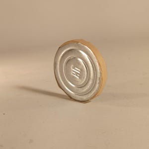 Op de afbeelding: Een zilveren metalen flesstopper met een cirkelvormig ontwerp en een bruine kartonnen rand. De stopper heeft een geribbeld oppervlak en een klein, in reliëf aangebracht ontwerp in het midden. Het object staat tegen een neutrale achtergrond.