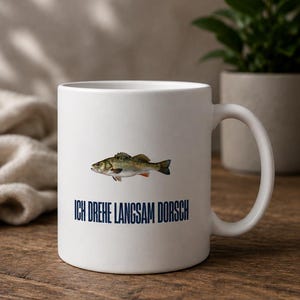 Könnte beinhalten: Ein weißer Keramikbecher mit einer detaillierten Fischillustration über dem dunkelblauen Schriftzug ICH DREHE LANGSAM DORSCH. Der Becher verfügt über einen Standardhenkel auf der rechten Seite und eine glatte, mattierte Außenfläche.
