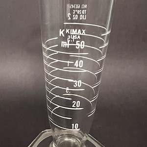 May include: A clear glass conical graduated cylinder with a wide mouth and a hexagonal base. White markings indicate measurements up to 50 ml. The cylinder has the text "KIMAX" and "USA" printed on it.