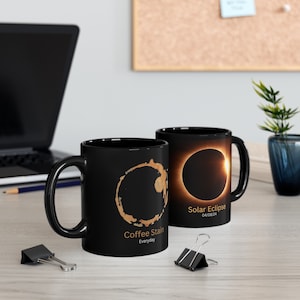 May include: Two black coffee mugs with different designs. One mug features a coffee stain with the text "Coffee Stain Everyday". The other mug features a photo of a solar eclipse with the text "Solar Eclipse 04/08/24".