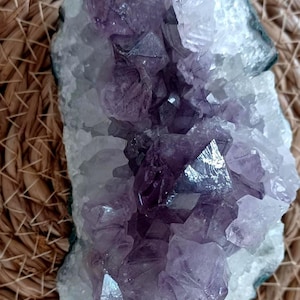 Peut inclure: Une géode d'améthyste naturelle avec une structure cristalline. La géode présente un amas de cristaux violets foncés entourés d'un extérieur blanc et rugueux. La géode est présentée sur une surface tissée brun clair.