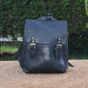 Può includere: Uno zaino in pelle nera con due grandi fibbie e una chiusura a lembo. Lo zaino ha un aspetto vissuto ed è realizzato in pelle di alta qualità.