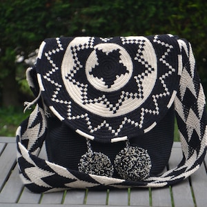 Puede incluir: Bolso de hombro de crochet negro y blanco con un patrón circular y dos pompones negros. El bolso tiene una correa larga con un patrón geométrico.