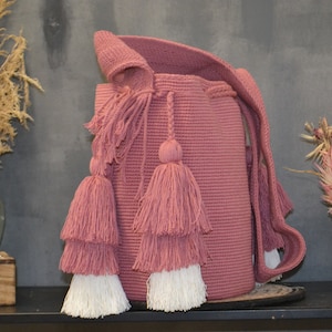 May include: A pink crocheted bag with white tassels. The bag has a long strap and is made of a soft, textured material.