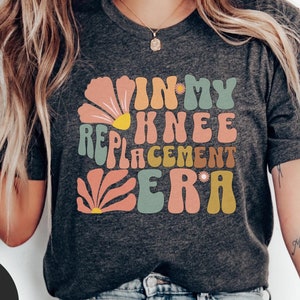 May include: A dark heather grey t-shirt with a retro-style graphic that says "In My Knee Replacement Era". The graphic features colorful flowers and swirls.