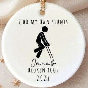 Op de afbeelding: Witte kerstbal van keramiek met zwarte tekst die "Ik doe mijn eigen stunts" zegt en een stick figure met krukken. De tekst "Jacob Broken Foot 2024" staat ook op de kerstbal.
