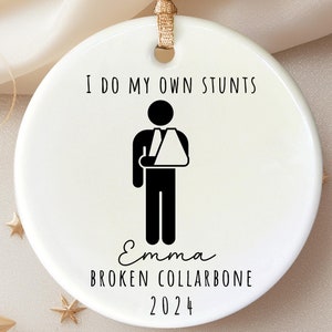 May include: White ceramic Christmas ornament with a black silhouette of a person with a broken arm. The ornament reads "I do my own stunts" and "Emma Broken Collarbone 2024".