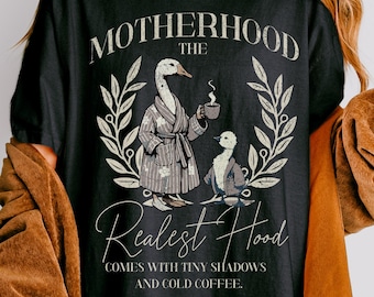 May include: Black graphic tee with the text "MOTHERHOOD The Realest Hood" and a goose illustration. One goose wears a robe and holds a coffee cup, the other is seated. The tee also includes the phrase "COMES WITH TINY SHADOWS AND COLD COFFEE."