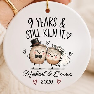 May include: A round white ceramic ornament featuring the text 9 YEARS & STILL KILN IT. The design includes two illustrated mugs, one wearing a top hat and bowtie, the other with a floral veil. The names Michael & Emma and 2026 are printed below.