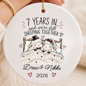 May include: A round white ceramic ornament featuring a cartoon illustration of two sheep in a bed. The text reads 7 YEARS IN... and we're still SHEEPING TOGETHER. Drew & Nikki 2026. The design includes small red hearts and a gold hanging string.