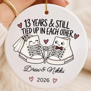 May include: A round white ceramic ornament featuring a graphic of two white trainers tied together with laces. The text reads 13 YEARS & STILL TIED UP IN EACH OTHER, Drew & Nikki, and 2026. The design includes small red hearts and a bridal veil.