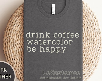 May include: Dark heather grey t-shirt with the text "drink coffee watercolor be happy" in white lettering. The shirt also has the text "LeCustomce DESIGNED BY JESS" in grey lettering.