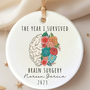 Peut inclure: Ornement en céramique blanche avec un motif floral coloré et le texte "The year I survived brain surgery Noreen Garcia 2023".