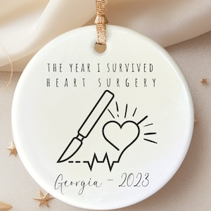May include: White ceramic ornament with black text and a graphic of a heart, a scalpel, and a heartbeat line. The text reads "The year I survived heart surgery Georgia - 2023".