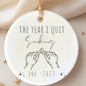 以下が含まれることがあります： 白いセラミック製のオーナメントで、黒い文字で「The Year I Quit Smoking」と書いてあり、2本の指でタバコを折る黒い線画が描かれています。オーナメントには「Joe - 2023」という文字と小さな星が2つ描かれています。