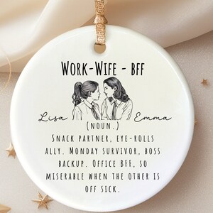 May include: White ceramic bauble featuring a black and white illustration of two women. The text reads "WORK-WIFE - BFF" and includes the names "Lisa" and "Emma". The text also reads "(NOUN.) SNACK PARTNER, EYE-ROLLS ALLY. MONDAY SURVIVOR, BOSS BACKUP. OFFICE BFF, SO MISERABLE WHEN THE OTHER IS OFF SICK."
