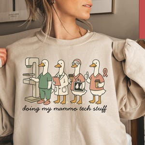 May include: A sand-colored sweatshirt with a graphic of four cartoon ducks in medical attire. The ducks are depicted with a mammogram machine, holding charts, coffee cups, and a pink ribbon. The text "doing my mammo tech stuff" is below the ducks.