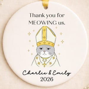 May include: A circular white ceramic ornament with a yellow ribbon. It features a cartoon cat in religious attire, with the text "Thank you for MEOWING us." and the names "Charlie & Emily 2026."