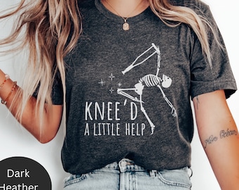 May include: A dark heather grey t-shirt with a white skeleton graphic and the text "Knee'd a little help".