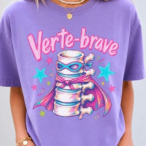 Może przedstawiać: Jasnofioletowy t-shirt z grafiką kręgosłupa w masce i pelerynie superbohatera. Napis "Verte-brave" jest nadrukowany na różowo nad ilustracją, która jest otoczona małymi różowymi, niebieskimi i żółtymi gwiazdkami.