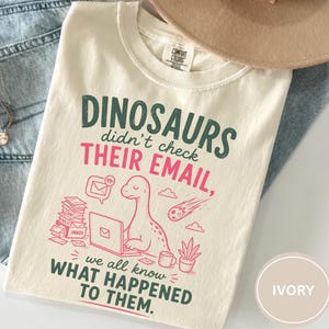 May include: An ivory t-shirt featuring a graphic print with dark green and pink text that reads: DINOSAURS didn't check THEIR EMAIL, we all know WHAT HAPPENED TO THEM. The design includes a dinosaur at a laptop, a stack of mail, and a meteor.