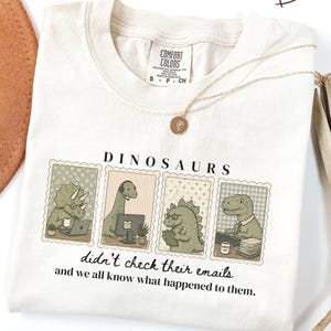 May include: An off-white t-shirt featuring a graphic of four dinosaurs in office settings with the text "DINOSAURS didn't check their emails and we all know what happened to them." The shirt has a Comfort Colors brand label at the inner neckline.