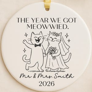 May include: A round, off-white ceramic ornament with a yellow ribbon. The ornament features a black and white illustration of two cats, one in a tuxedo and the other in a wedding dress. Text reads "THE YEAR WE GOT MEOWWIED" and "Mr & Mrs. Smith 2026".