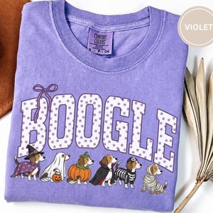 Puede incluir: Una camiseta de cuello redondo color violeta con acabado lavado vintage. El frente presenta el texto BOOGLE en blanco con lunares violetas y un lazo violeta. Debajo, seis perros beagle están ilustrados con varios disfraces de Halloween: bruja, fantasma, calabaza, vampiro, esqueleto y momia.