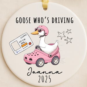 May include: A circular, cream-coloured ornament featuring a cartoon goose in a pink helmet and sunglasses, driving a pink car-shaped shoe. The ornament has the text "GOOSE WHO'S DRIVING" and "Joanna 2025". A driving license and stars are also depicted.