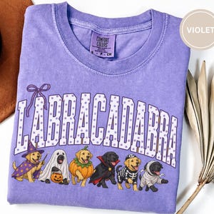 May include: A violet t-shirt featuring the text LABRACADABRA in white with purple polka dots. Below the text are six Labrador dogs in Halloween costumes: a wizard, a ghost, a pumpkin, a vampire, a skeleton, and a mummy. The shirt has a crew neck.